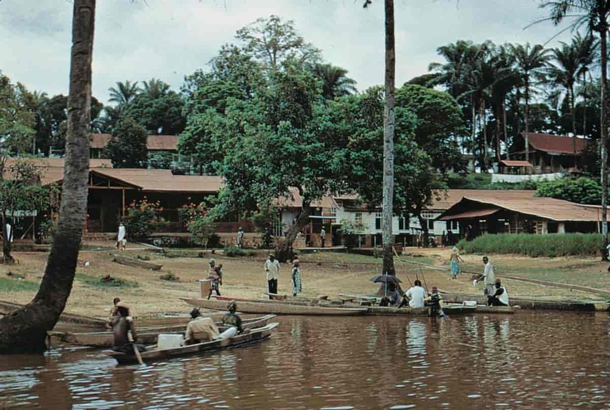 mennesker i lave, avlange båter på en brun elv. lave hus og trær i bakgrunnen. foto.