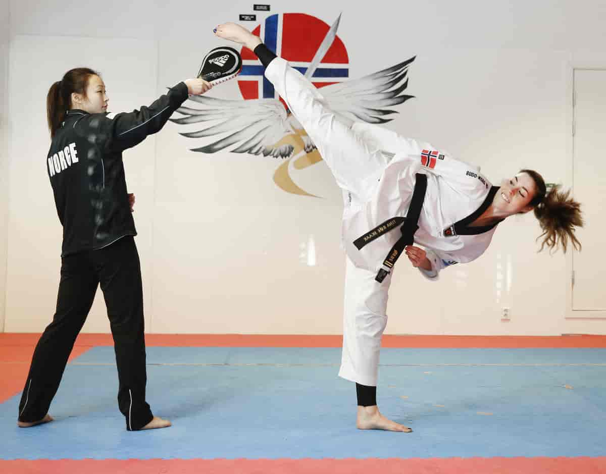 Foto av Røe Skaar i taekwondodrakt med svart belte som sparker mot en liten plate en person holder opp i lufta.