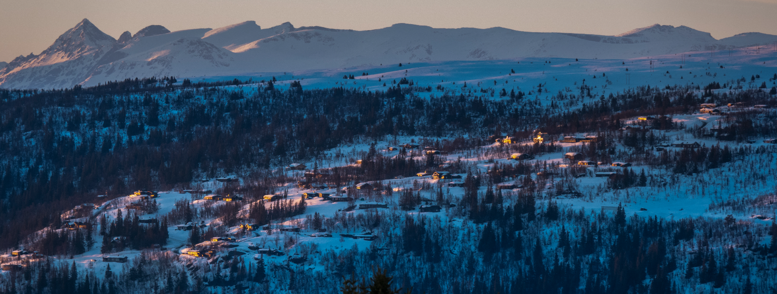 Foto av et fjellterreng med masse hytter. I bakgrunnen kan man se fjellet Jotunheimen. Det er vinter og mye snø. 