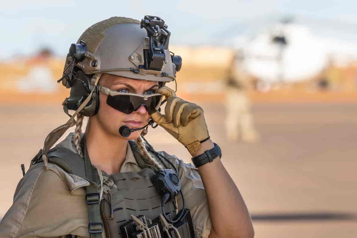 Foto av en kvinne i sandfarget uniform med skuddsikker vest, hansker og hjelm med øreklokker og mikrofon. Hun har et par solbriller på som hun trekker ned så hun kikker over kanten.