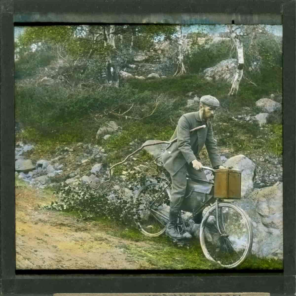 Håndkolorert bilde av Anders Beer Wilse med sitt kamerautstyr syklende fra Filefjell til Lærdal i 1902.