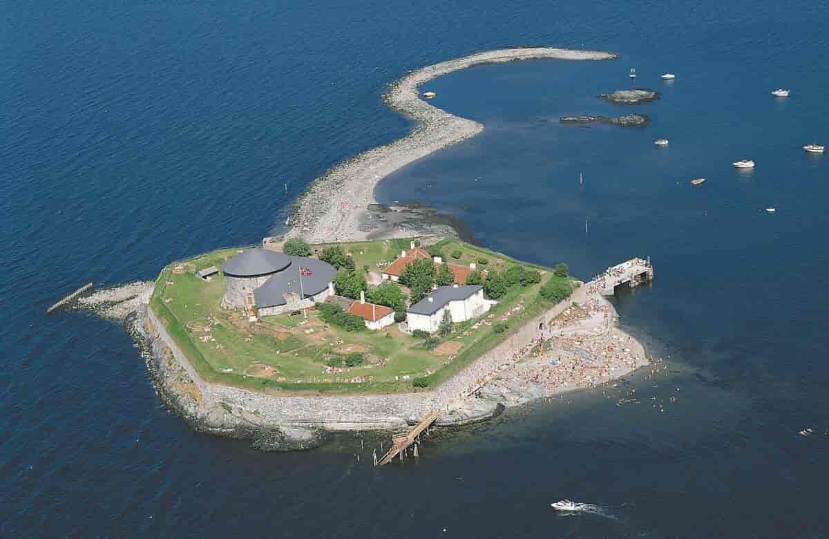Flere mindre bygninger på en liten øy med hav rundt hele øya. Midt på øya ligger det en rund bygning i stein med grått tak. En vei går et stykke ut i havet fra øya.