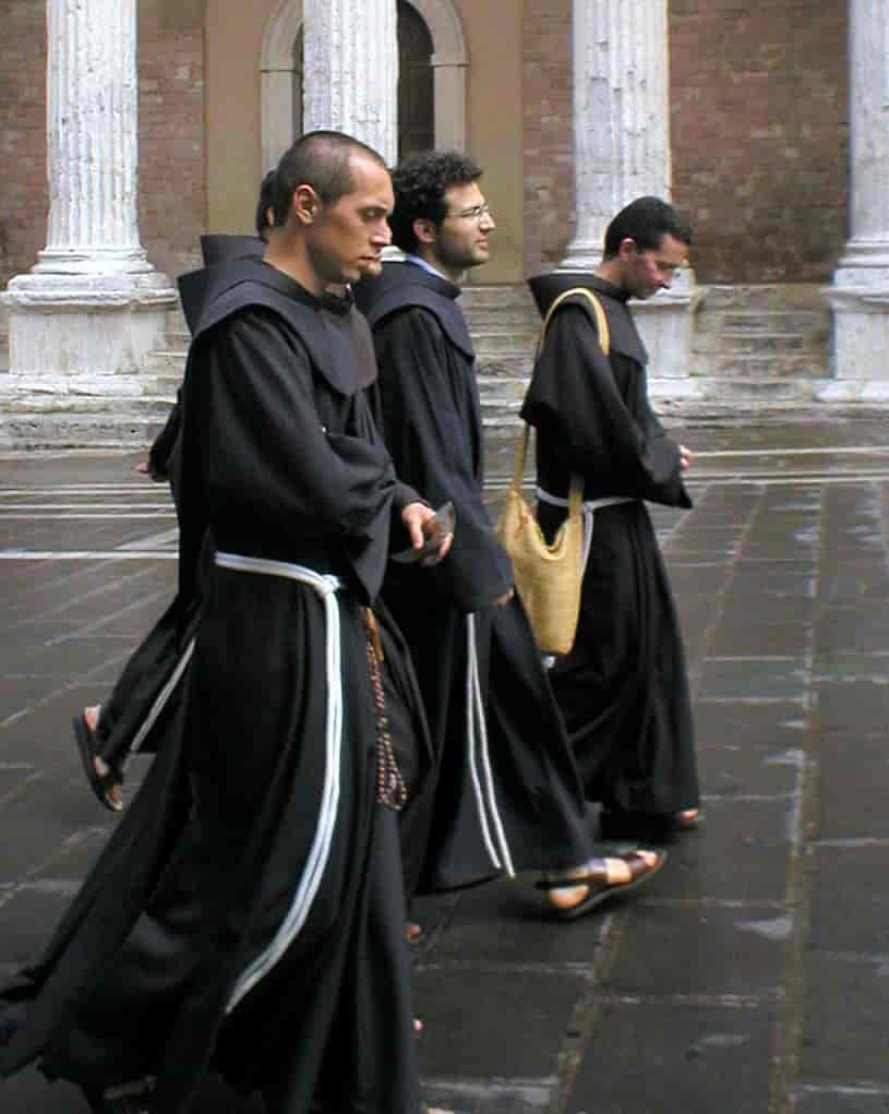 Foto av fire menn i lange, svarte kapper med hvite snorer som belter rundt livet som går over en steinlagt plass. De går i sandaler, og en av mennene har en rosenkrans hengende i beltet.