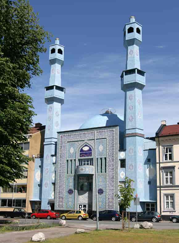 Fotografi av en lyseblå moské med to tårn. Den er dekket av mosaikk, og den har en kuppel på taket. Den ligger ved en gate mellom to hus som ser ut som bygårder med leiligheter.