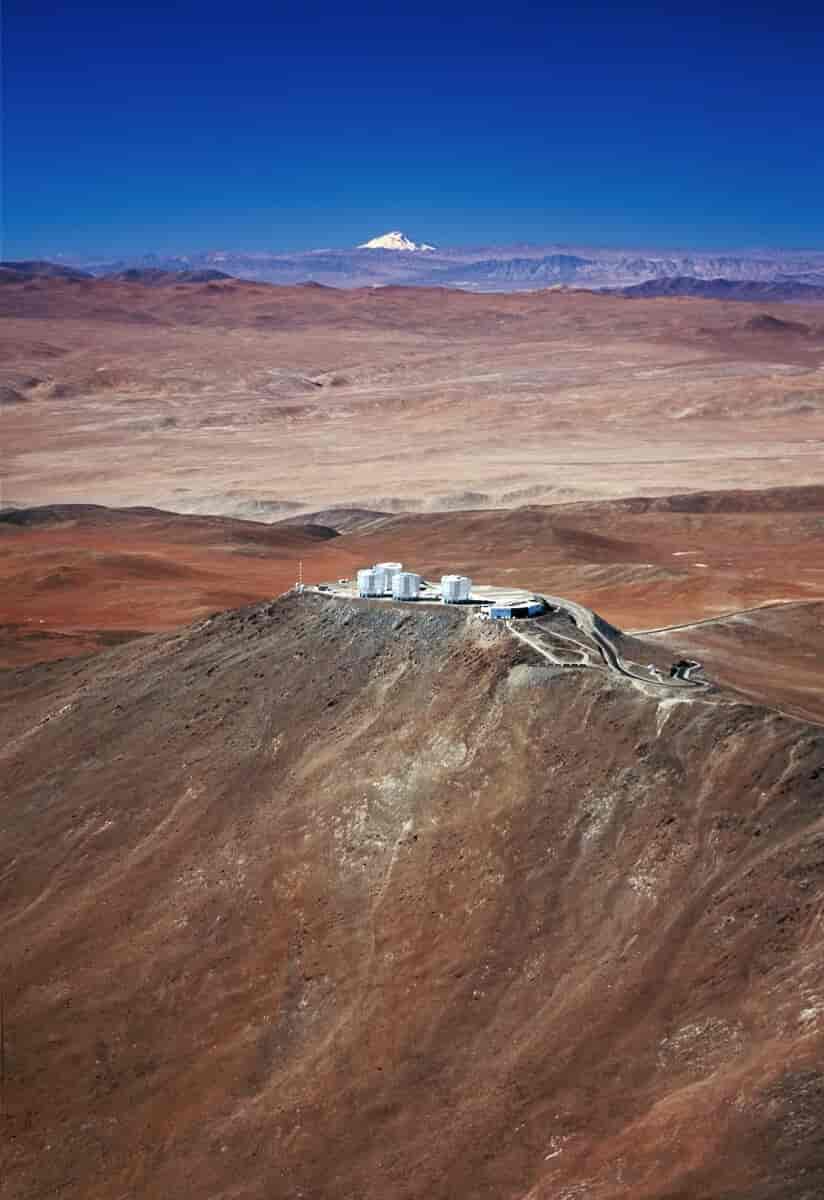 Observatoriet består av fire hvit sylinderformede bygninger. Man kan skimte en mast og noen lave bygninger. Observatoriet ligger på toppen av et brunt fjell, med øde, røde fjell i bakgrunnen. Det går flere veier opp til observatoriet.