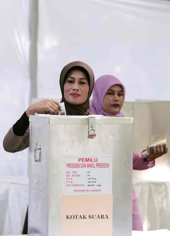 Foto av to kvinner og en valgurne i metall. Den ene kvinnen er i ferd med å putte en stemmeseddel i valgurnen.
