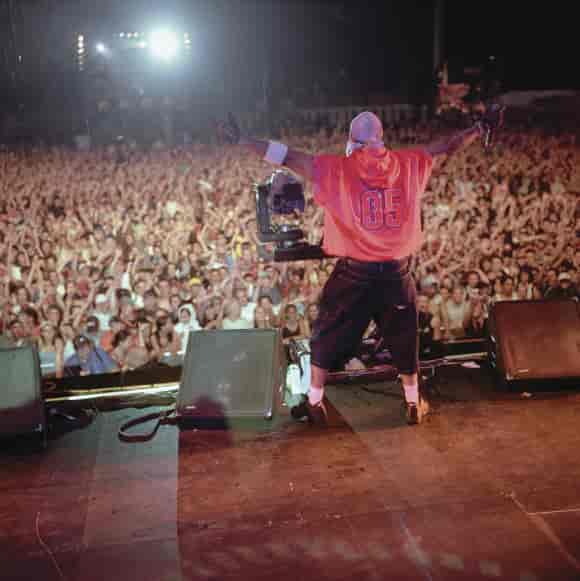 En musiker i store klær står med armene ut til siden på en scene. Foran ham er det et hav av publikum.
