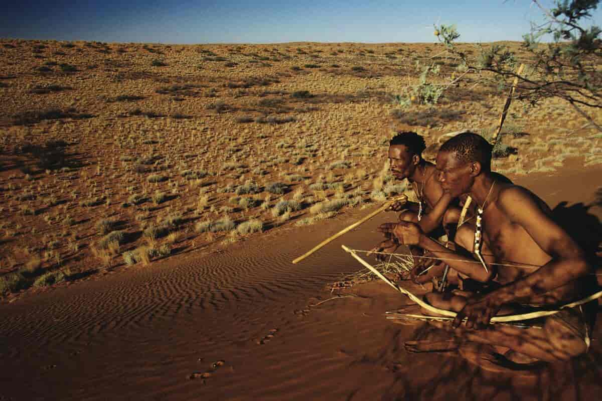 To menn med pil og bue sitter på huk i et ørkenlandskap. Foto.