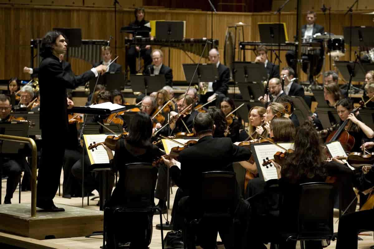 Bildet viser en dirigent som står foran et orkester. Han holder en taktstokk i hånden og musikerne følger oppmerksomt med på dirigenten. man ser først og fremst strykere, og kan skimte blåsere lenger bak i bildet.