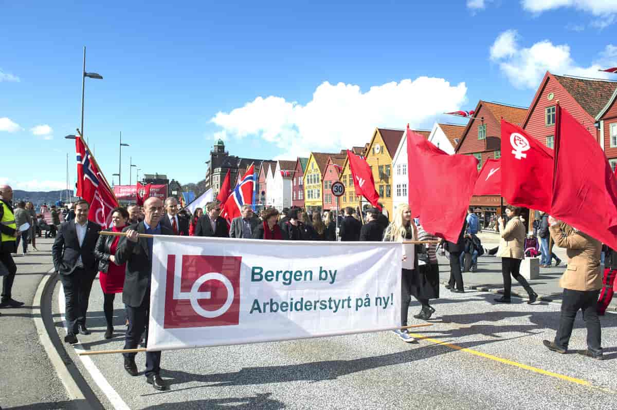Foto av 1. mai-tog med røde faner og norske flagg. Forrest går to personer med et banner hvor det står «LO. Bergen by Arbeiderstyrt på ny!»