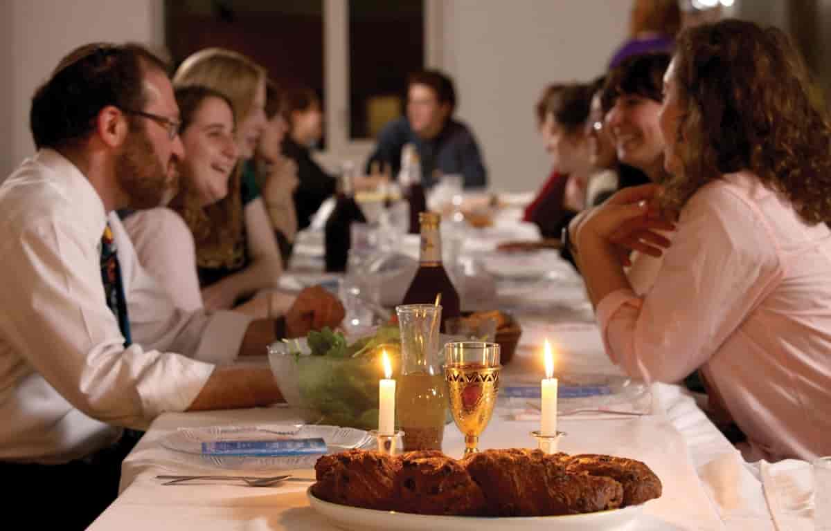 Voksne kvinner og menn sitter rundt et dekket bord. Det er brød og lys foran på bildet. En av mennene har en kalott på hodet.