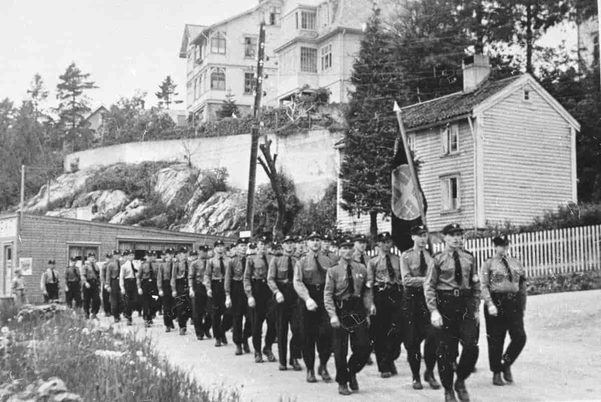 Svart-hvittfoto av om lag 50 uniformerte menn som marsjerer to i rekke. Mannen på rad to bærer et stort flagg med et emblem. I bakgrunnen to hus, en stor sveitservilla og et mindre trehus. 