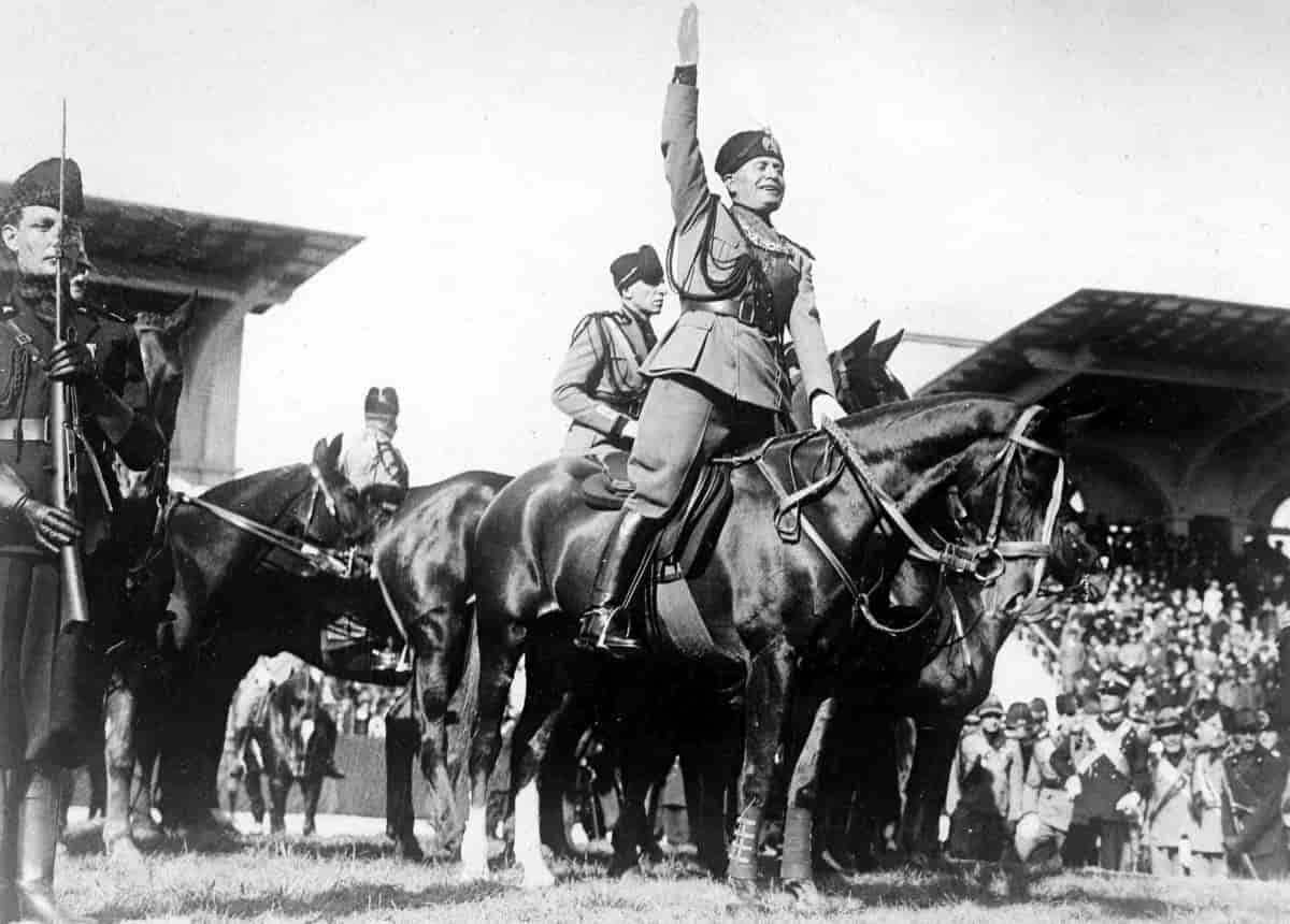 Svart-hvittfoto av uniformskledd Benito Mussolini. Han sitter på en svart hest, og har armen løftet rett opp. Rundt ham er det flere svarte hester med uniformskledde menn. En mann med uniform og gevær til venstre i bildet. I bakgrunnen masse mennesker på en tribune. 