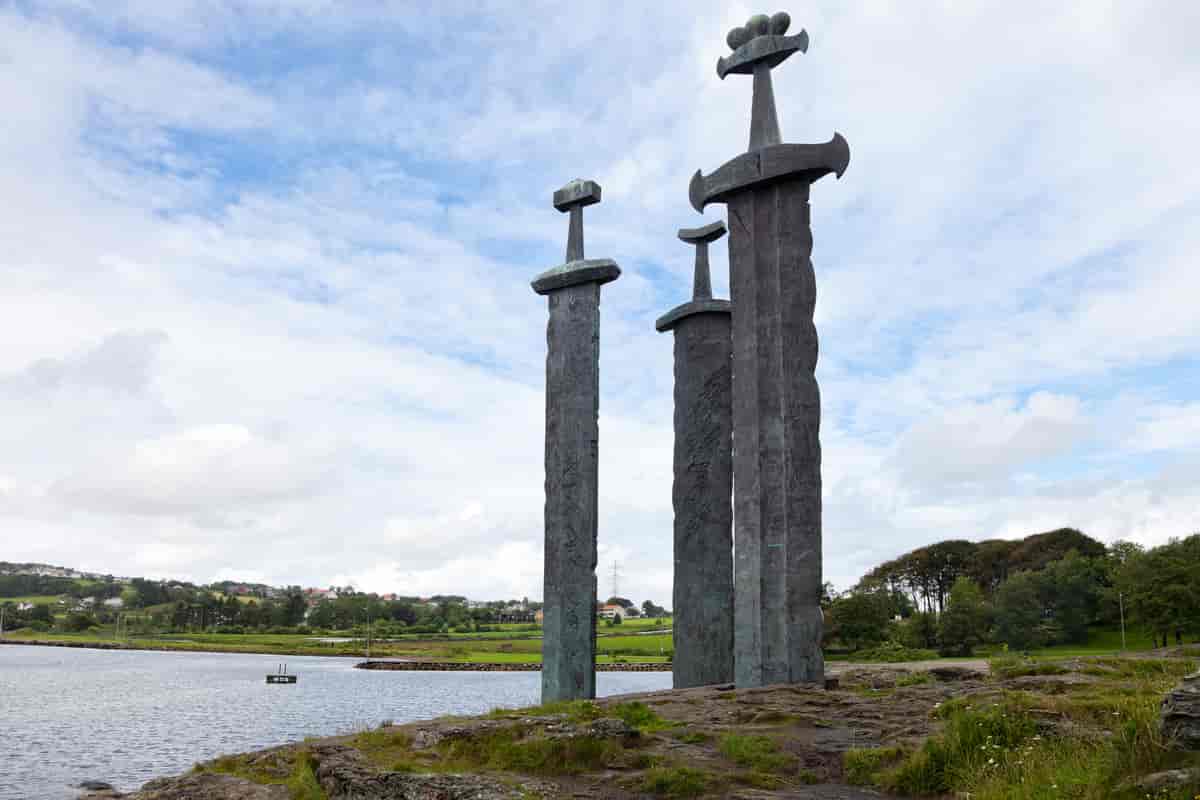 Foto av ein skulptur som ser ut som tre veldig store sverd som er stukke ned i bakken like ved sjøen