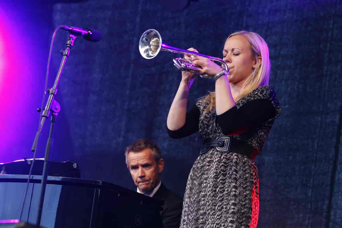 En lyshåret kvinne spiller konsentrert på en trompet. Bak henne sitter det en mann og spiller piano.