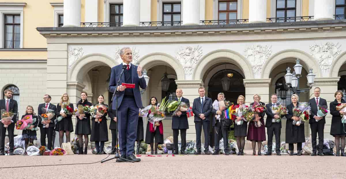 Foto av regjeringsmedlemmer på slottsplassen i Oslo. De fleste har blomsterbuketter i hendene. En har et norsk flagg, og en har et pride-flagg. Støre står litt foran de andre og snakker i mikrofon.