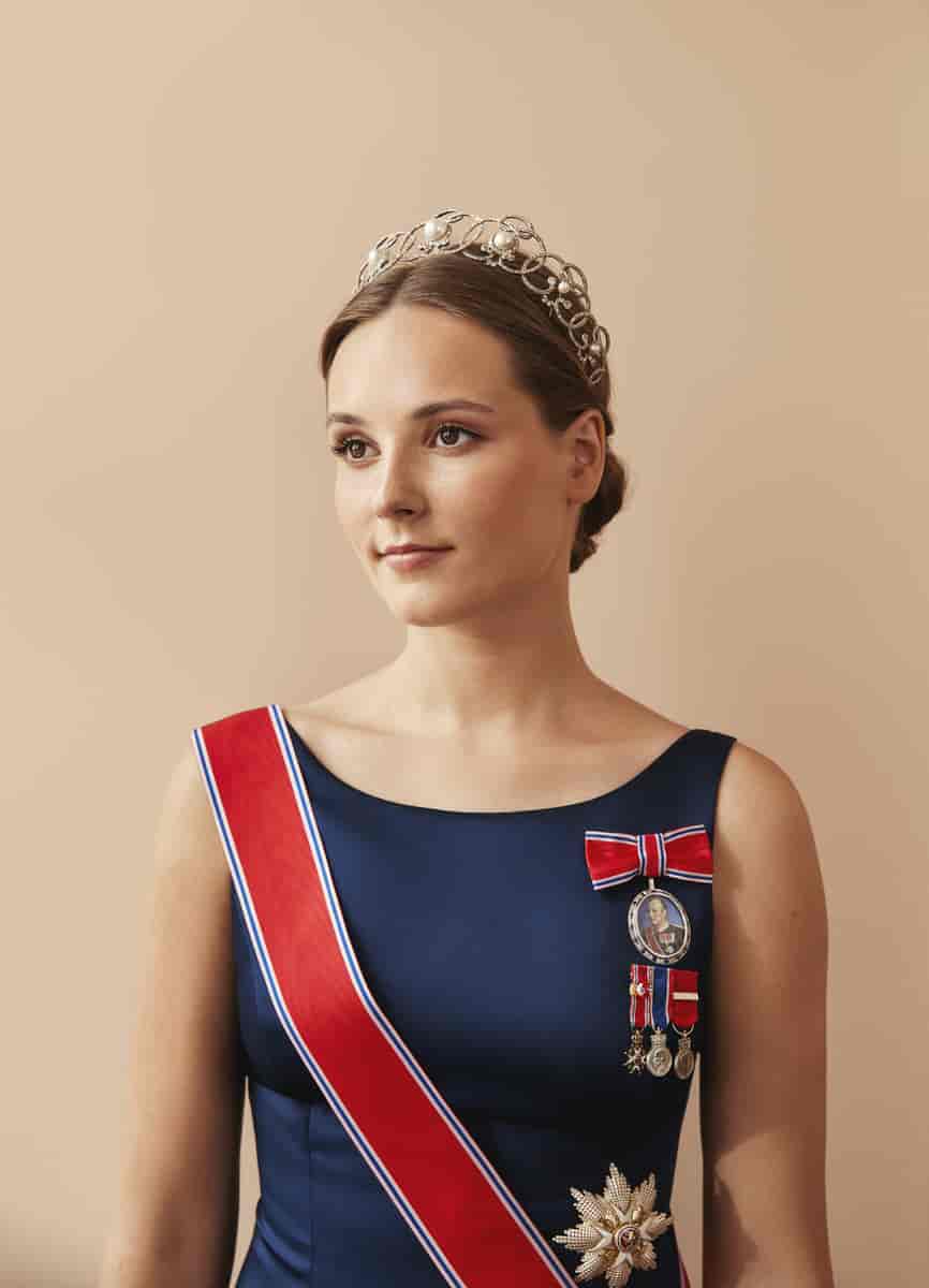 Portrett av en kvinne med en tiara med perler, rødt ordensbånd over en mørkeblå kjole og en medaljong med bilde av kong Harald 5. på brystet. Foto
