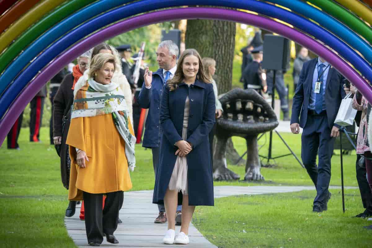 Foto av dronning Sonja og prinsesse Ingrid Alexandra går på en gangvei i slottsparken på vei under en regnbueskulptur. I bakgrunnen er en skulptur av en stor munn med føtter.