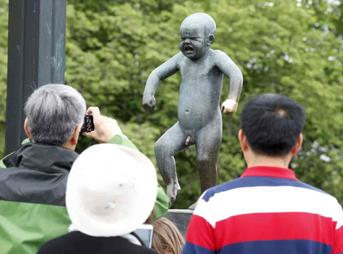 En bronsestatue av en liten, naken gutt som står på ett bein. Han har sammenknepne øyne og åpen munn. Foran statuen står tre personer. En av dem tar bilde med et kamera.