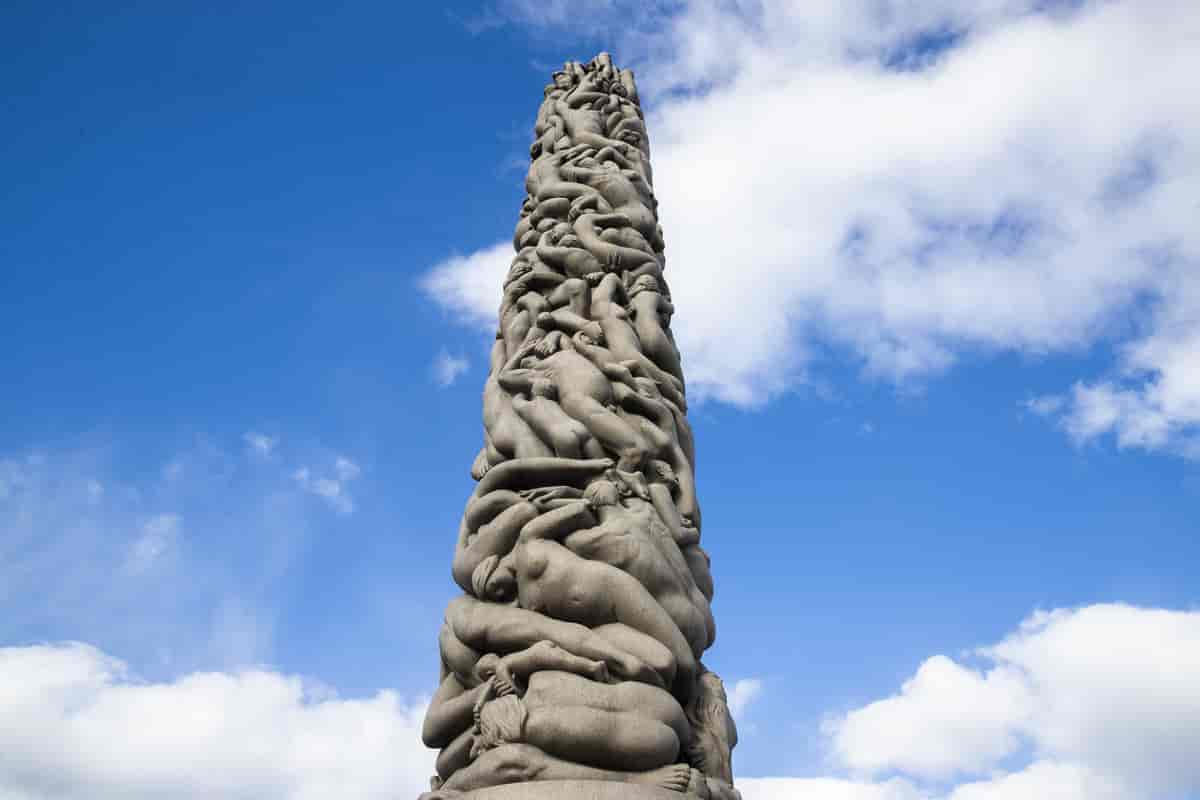 Skulptur i stein med form som en høy søyle. Oppover søylen klynger nakne mennesker seg sammen.