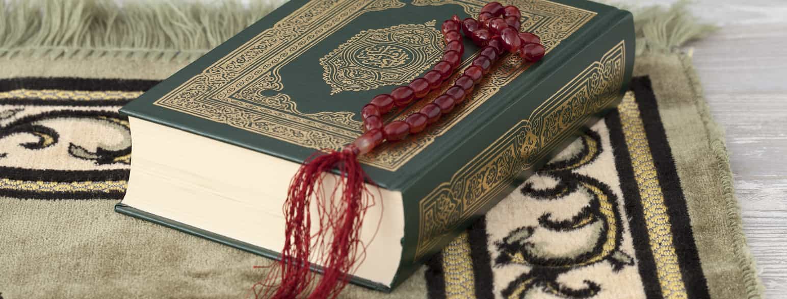 Fotografi av en Koran som ligger på et mønstret teppe. Koranen er en tykk, grønn bok som er dekorert med gullmønster. Oppå Koranen ligger et kjede av røde perler med en lang, rød dusk i enden.