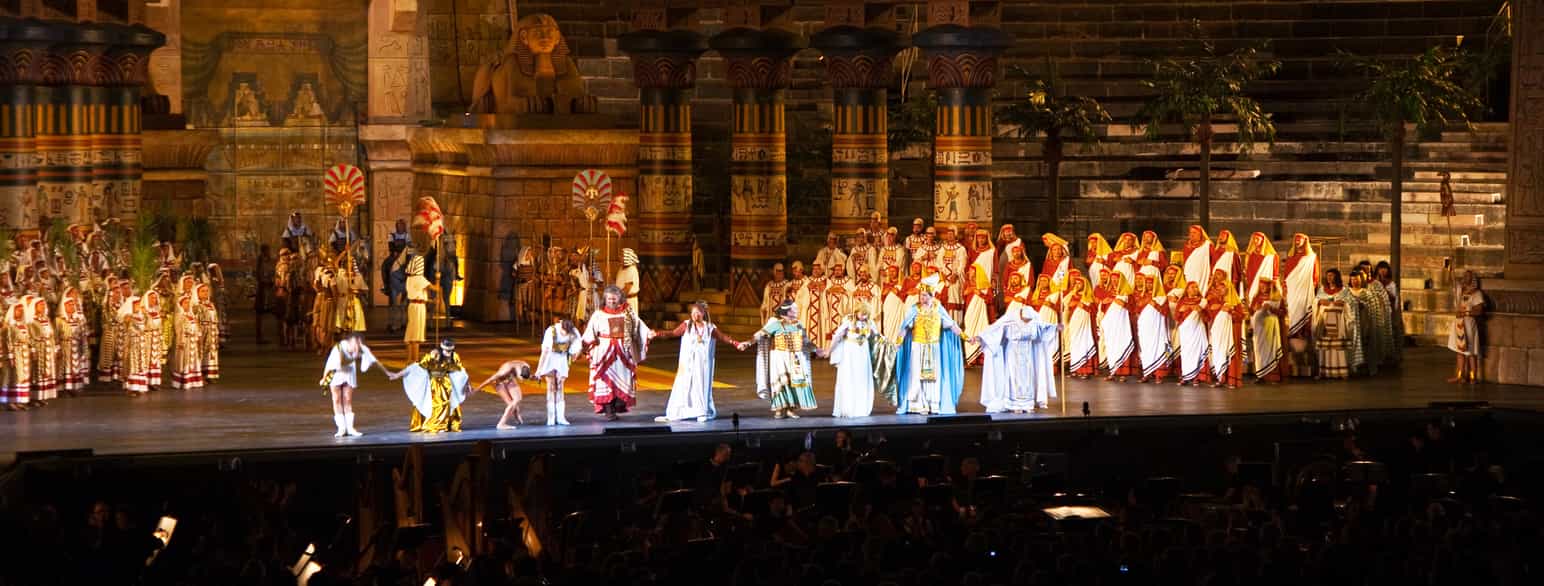 Fotografiet viser mange mennesker på en scene. De har på seg kostymer som viser at operaen foregår i det gamle Egypt.