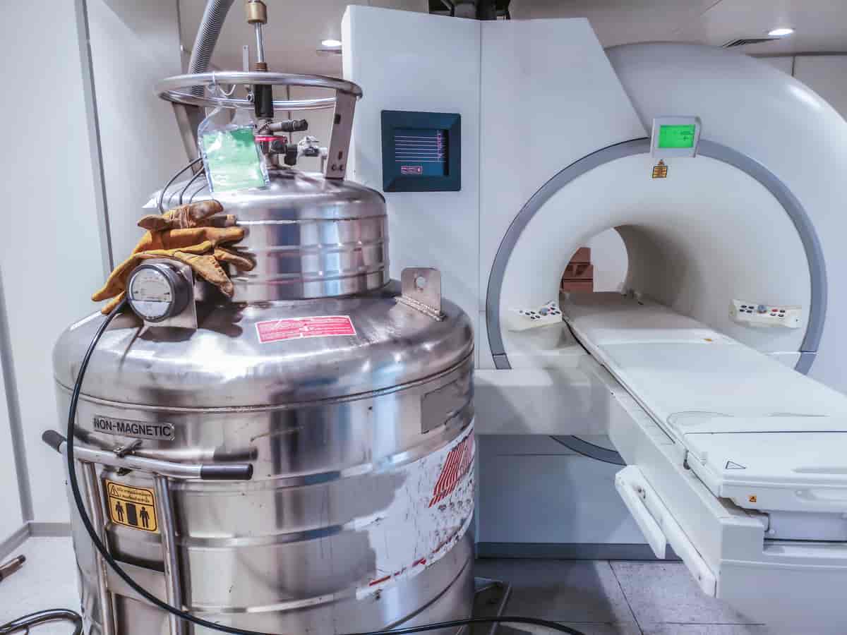 En MR-maskin er en stor hvit maskin med et hull i midten og en benk pasienten skal ligge på. Foran maskinen står det en stor tank i metall.