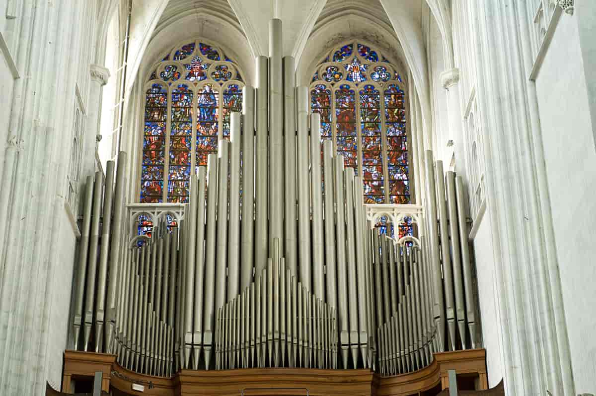 Et stort kirkeorgel med fargerike glassmalerier i bakgrunnen. Orgelet består av flere små og store rør som ser ut som piper som står rett opp.