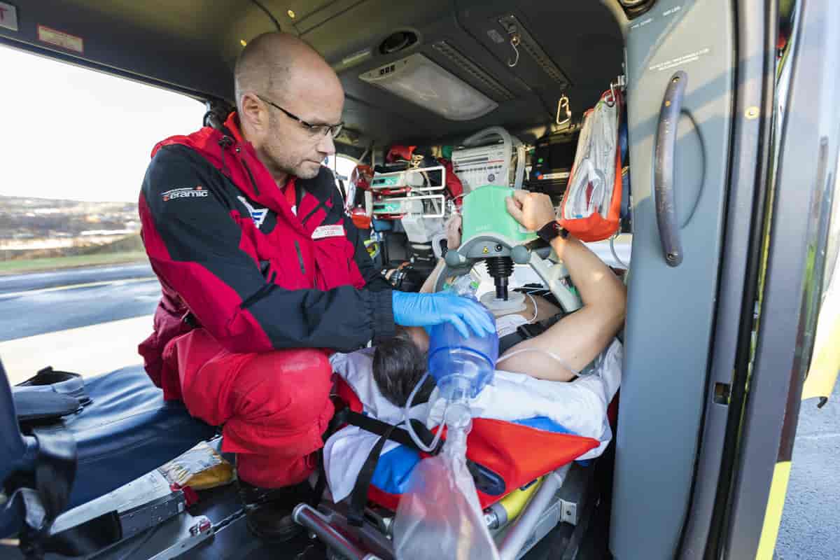 Pasienten ligger på en båre. Legen har på seg en rød ytterdress som uniform og bruker hansker. Han klemmer på en ballong koblet til en maske, for å hjelpe pasienten å puste.