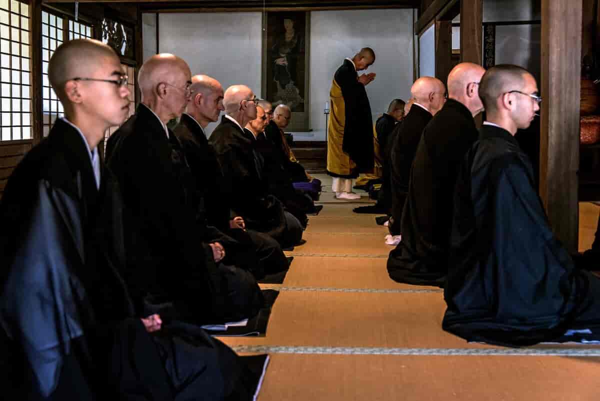 Fotografi av munker som kneler på et gulv. Munkene har svarte klær og glattbarberte hoder. De kneler i to rekker.