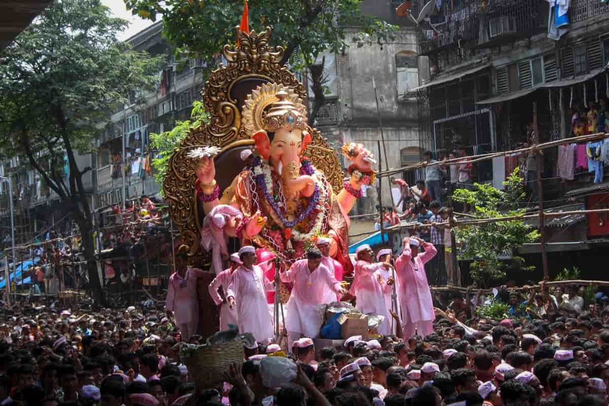 Fotograf av et opptog med veldig mange mennesker. Midt i bildet er en enorm, gullfarget trone med en figur av guden Ganesh. Ganesh har elefanthode. Han holder blomster i hendene og har mange blomsterkranser rundt halsen. Rundt tronen er det en gruppe folk med rosa klær. 