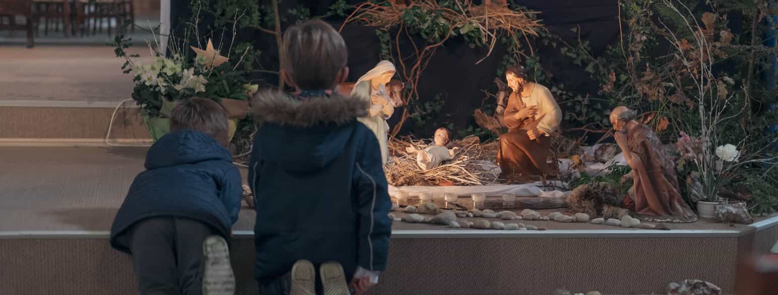 Fotografi av to barn som står med ryggen til kamera og ser på en julekrybbe. Julekrybben er figurer av Josef, Maria og et lite barn som ligger på en haug med strå. Bak Maria og Josef er det en figur av en ku og en av et esel. Til høyre kneler en annen figur.