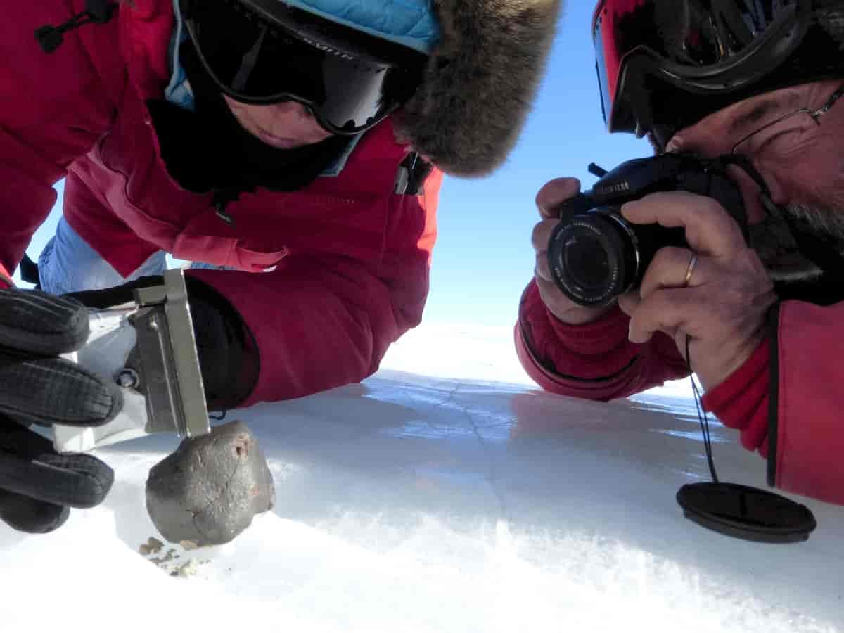 Nærbilde av to menn i røde jakker som ligger på isen. Mannen til venstre holder et måleinstrument som måler en svart stein. Mannen til høyre holder et kamera og tar bilde av steinen.