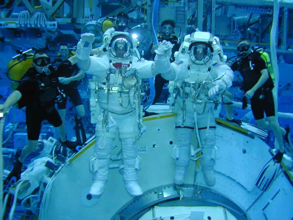 To astronauter i hvite romdrakter som ser ut som de svever i luften foran en romkapsel. I bakgrunnen er det fem mennesker i dykkerdrakter