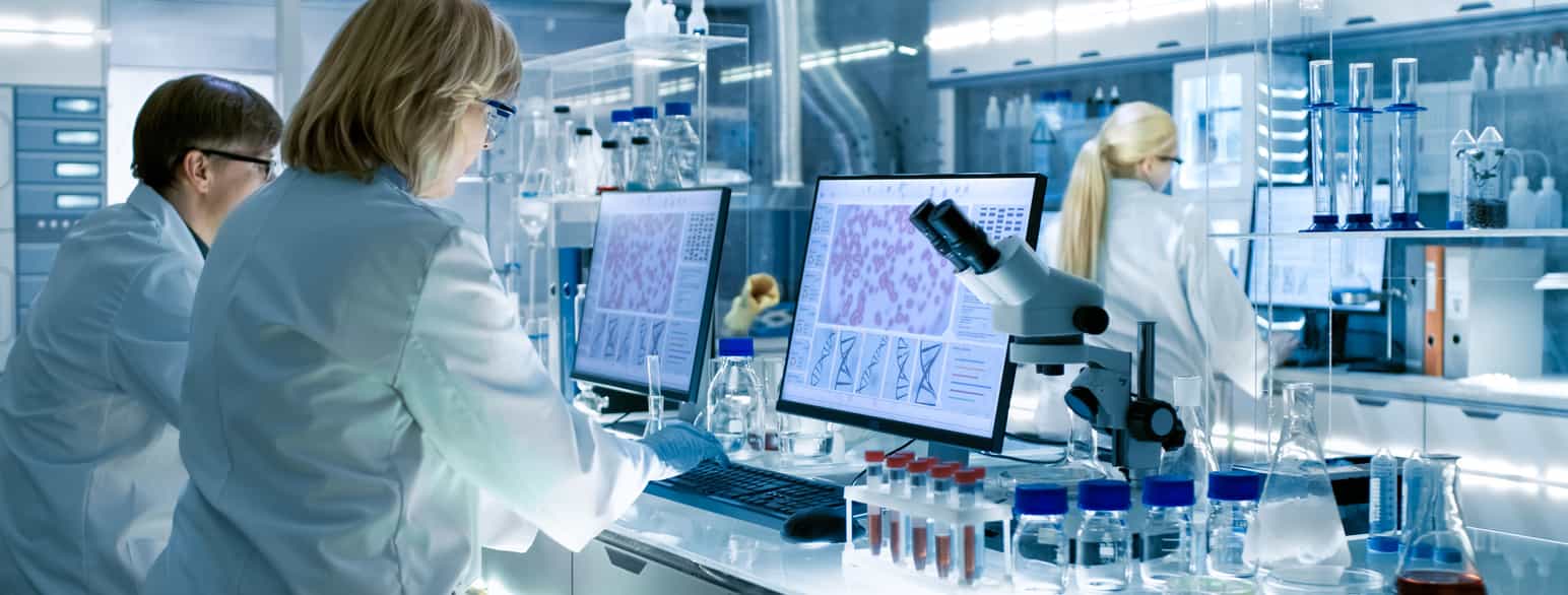 To forskere i en medisinsk lab. De har hvite frakker og på bordet deres er det mikroskop, prøverør og dataskjermer med store mikroskopbilder. 