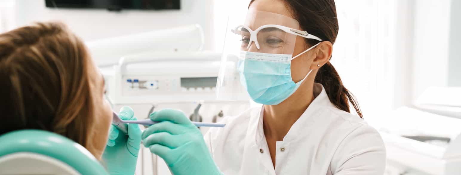 Tannlege med munnbind, plasthansker og skjold foran øynene ser inn i munnen til kvinne. Tannlegen bruker en pirkepinne av metall og et speil. 
