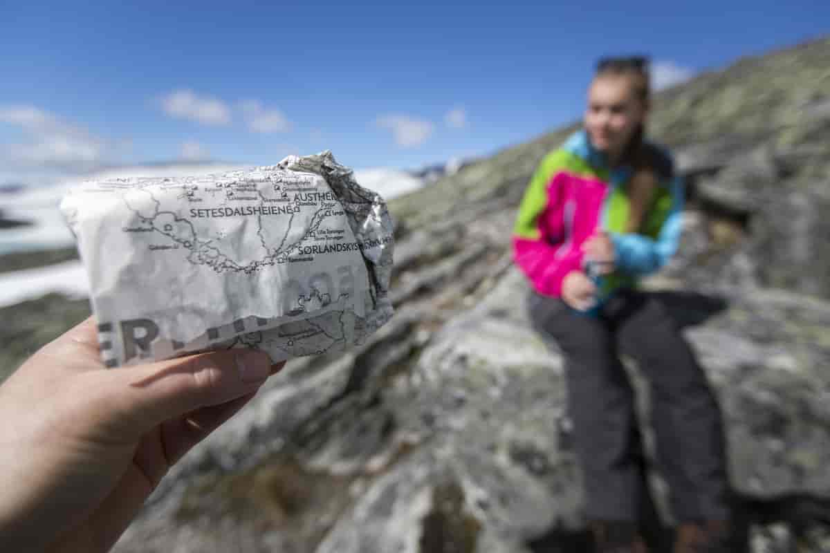 En hånd holder en matpakke, i bakgrunnen sees en person som sitter på en stor stein i et fjellandskap. Foto