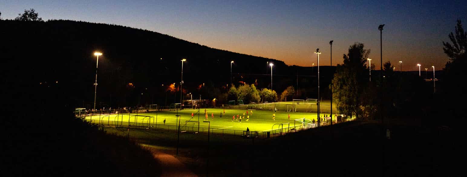 Foto av en fotballbane med gress opplyst av lyskastere. Mørk høstkveld i Norge.