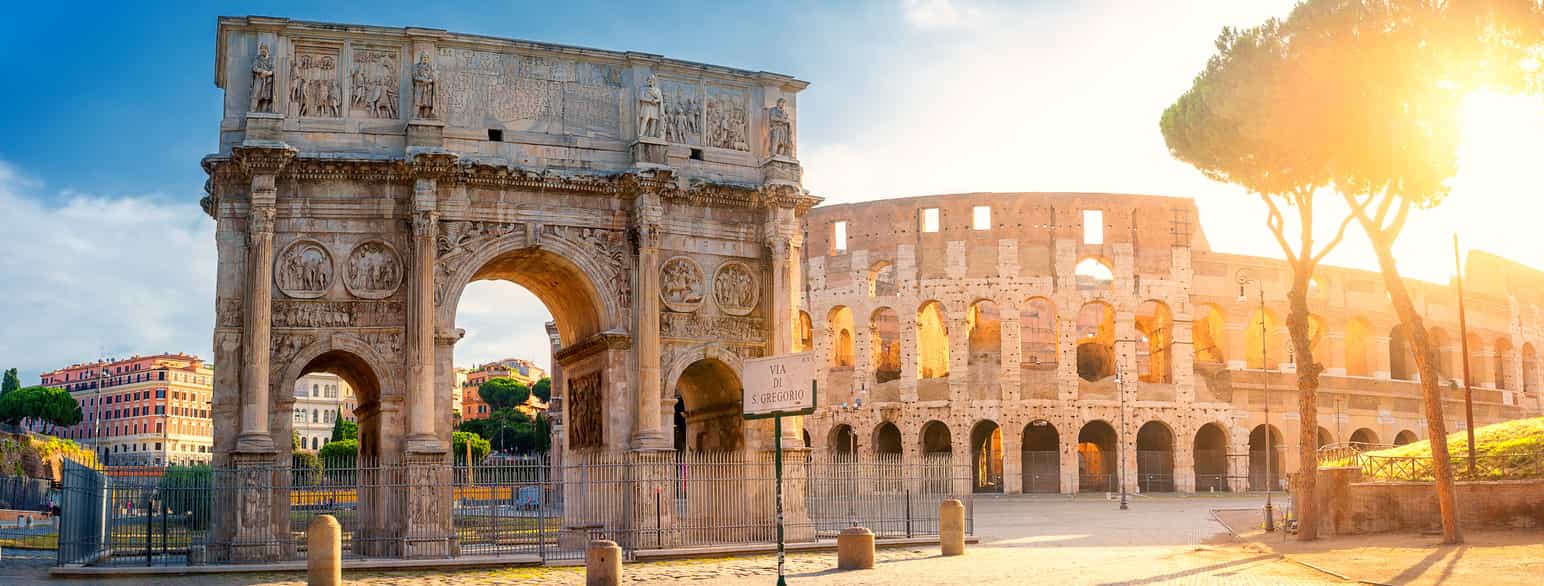 En triumfbue med en stor buet åpning i midten, og en liten buet åpning på hver side. I bakgrunnen ligger ruinene av et romersk amfiteater. Foto