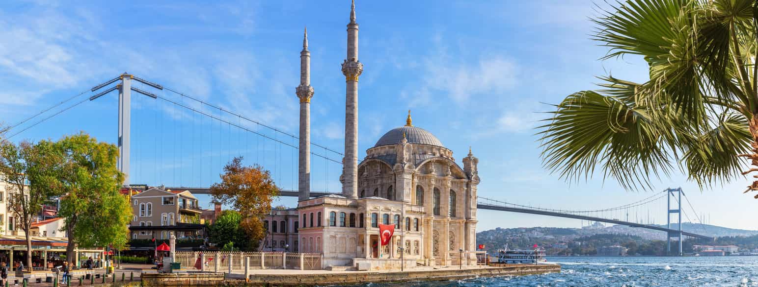En moské med hvelvet tak og to høye runde tårn rett ved vannet. I bakgrunnen en lang hengebro. På veggen av moskéen midt i bildet, henger et tyrkisk flagg. Foto