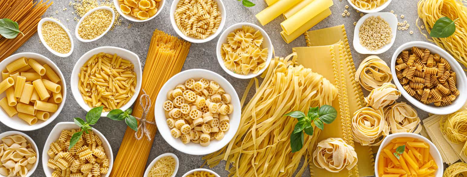 Forskjellige former for tørket pasta på en lys grå bordplate, blant annet spagetti, tagliatelle, penne, skruer, farfalle og makaroni. Noen få basilikumblader til pynt. Foto