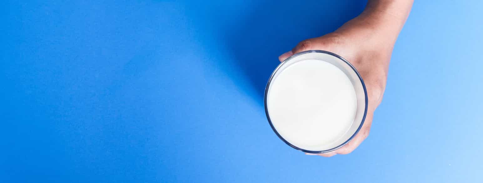 Bilde tatt rett ovenfra. En hånd holder rundt et glass melk som står på en blå bakgrunn. Foto