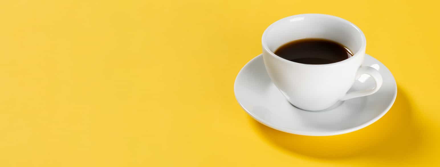 En hvit kaffekopp med skål under og kaffe oppi på gul bakgrunn. Foto