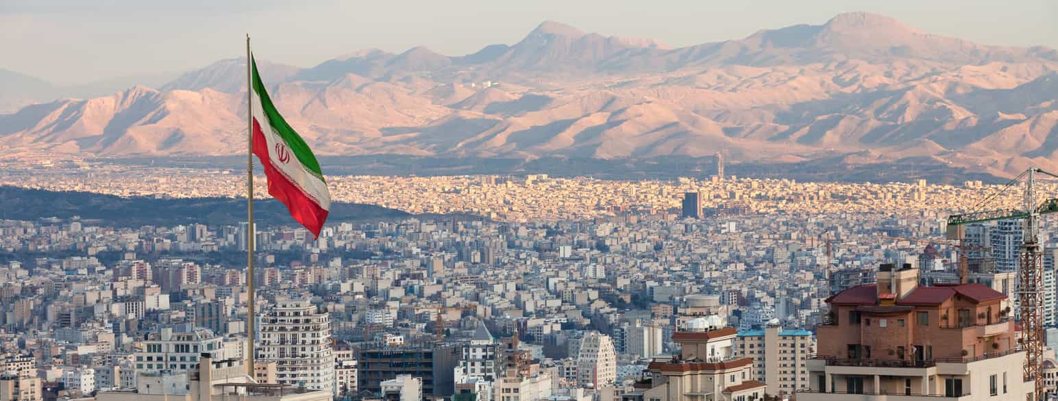 Luftfoto av en storby med høye fjell i bakgrunnen. Til venstre i bildet er en flaggstang med et iransk flagg. Foto