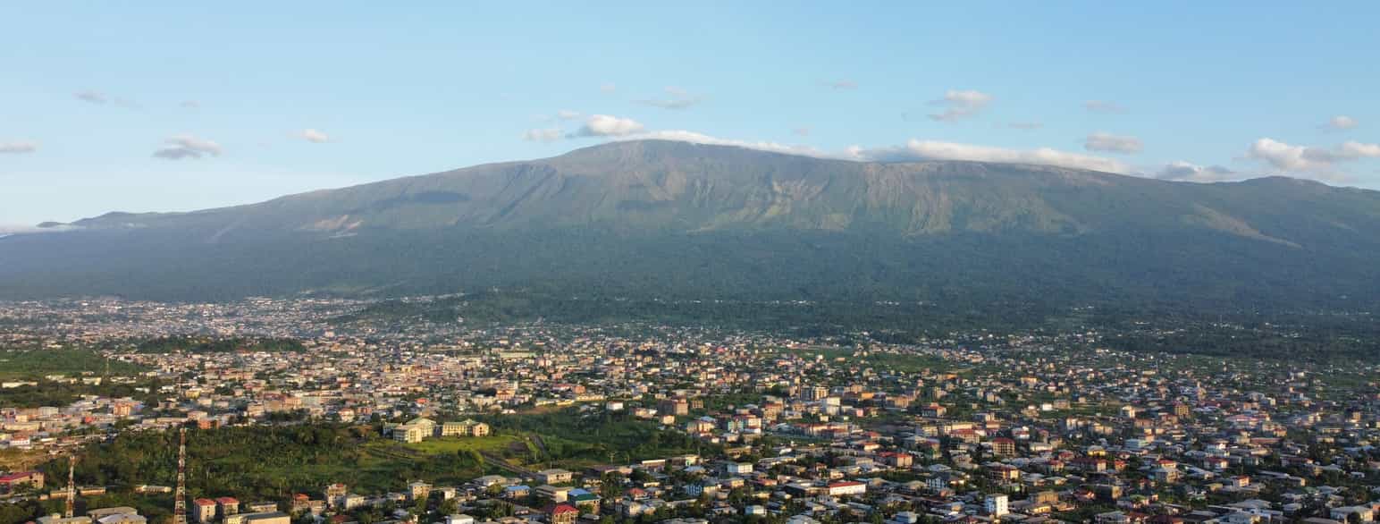 Oversiktsbilde av en by med grønne områder og et høyt fjell i bakgrunnen. Foto