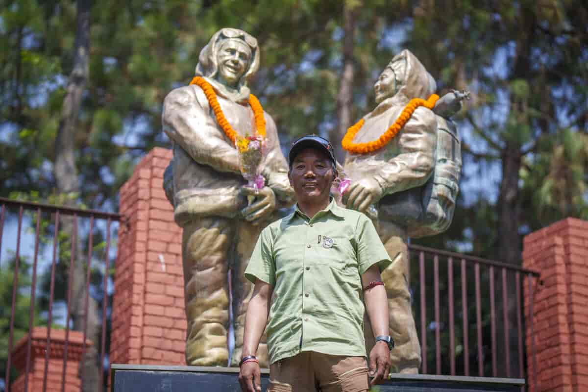 En mann i grønn skjorte  og blå caps står foran en statue av to menn. Mennene i statuen har oransje blomsterkranser rundt halsen. Foto.