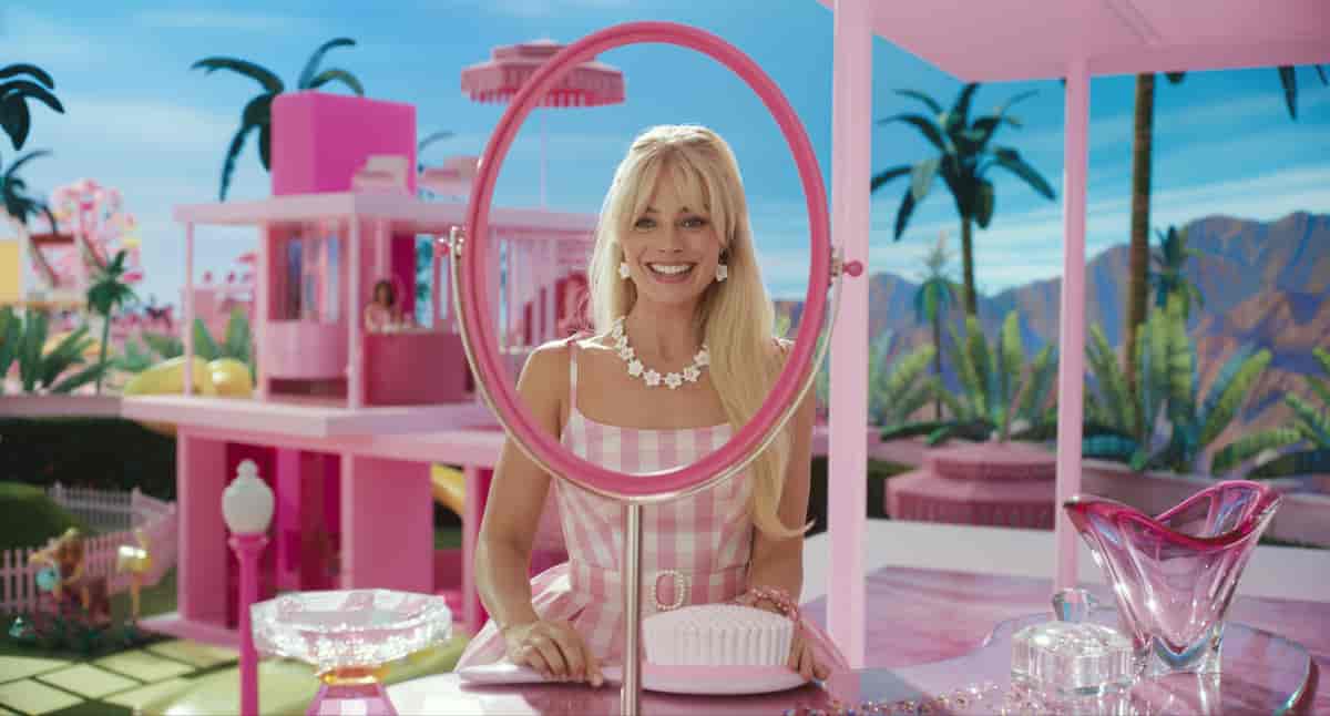 En kvinne med blondt hår og rosarutet kjole står ved en slags bardisk, omgitt av rosa effekter. I bakgrunnen et stort rosa Barbie-hus. Foto