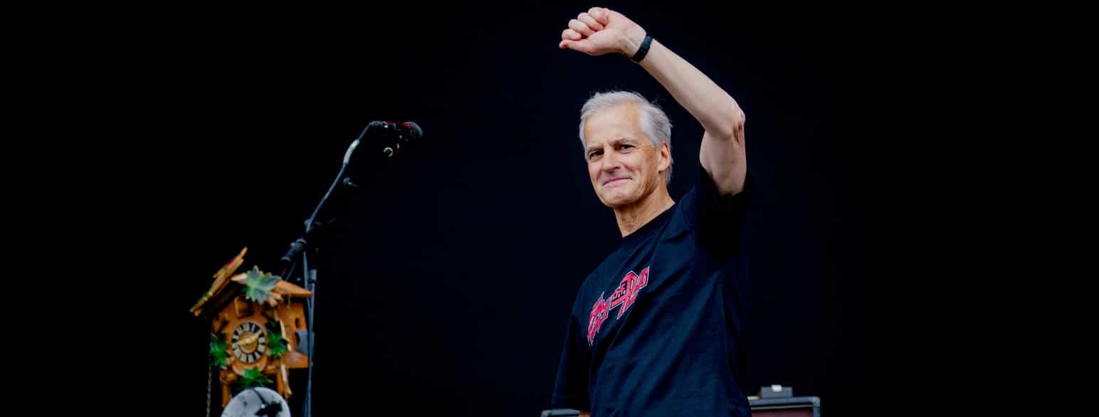 Foto av Støre i svart t-skjorte med rød tekst. Han løfter venstre arm i været med knyttet neve. Til venstre i bildet er en mikrofon på stativ og et gjøkur. Bakgrunnen er helt svart.