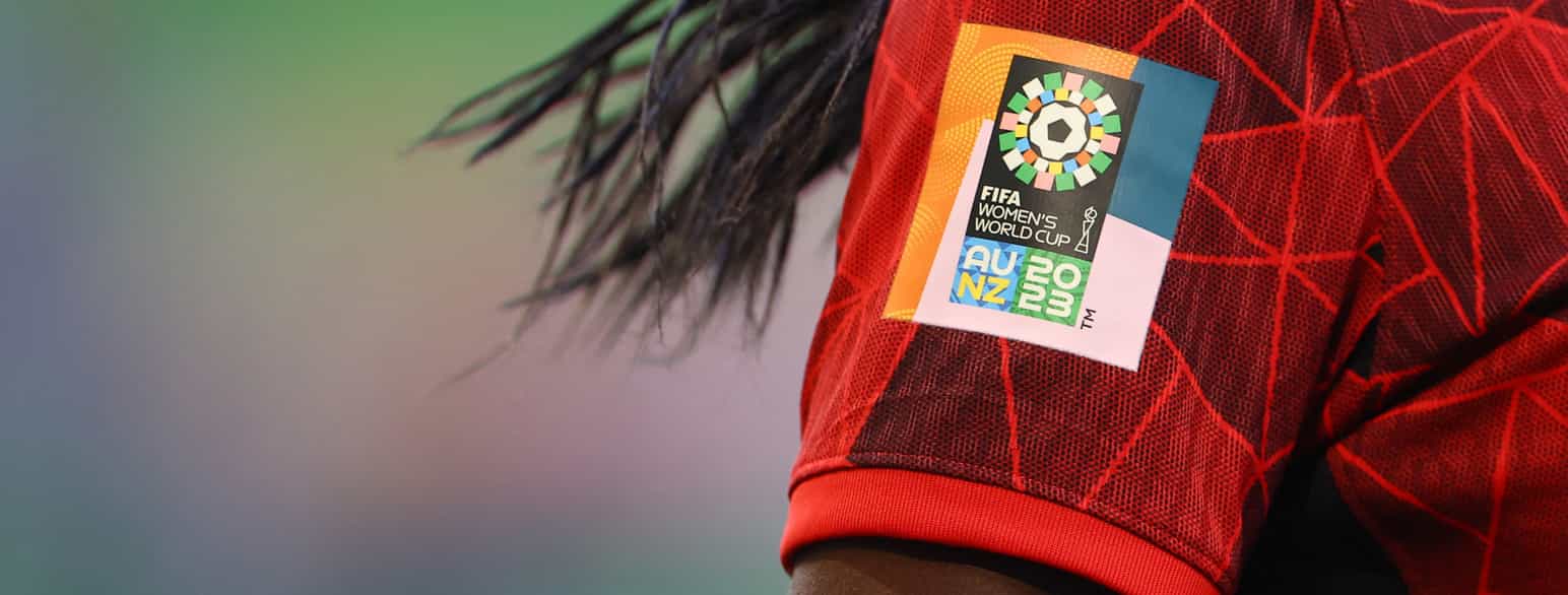 Foto av skulderen og overarmen til en person med rød fotballtrøye og langt svart hår. På ermet er et merke med logoen til fotball-VM 2023.