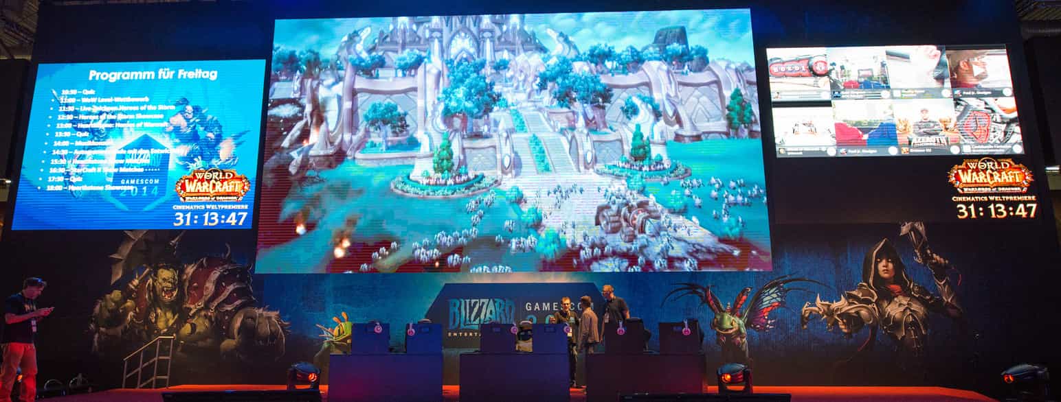 Tre skjermer henger over en stand i en messehall. Den største skjermen i midten viser spillet World of Warcraft. Under skjermene står noen mennesker og man ser logoen til Blizzard Entertainment. Foto